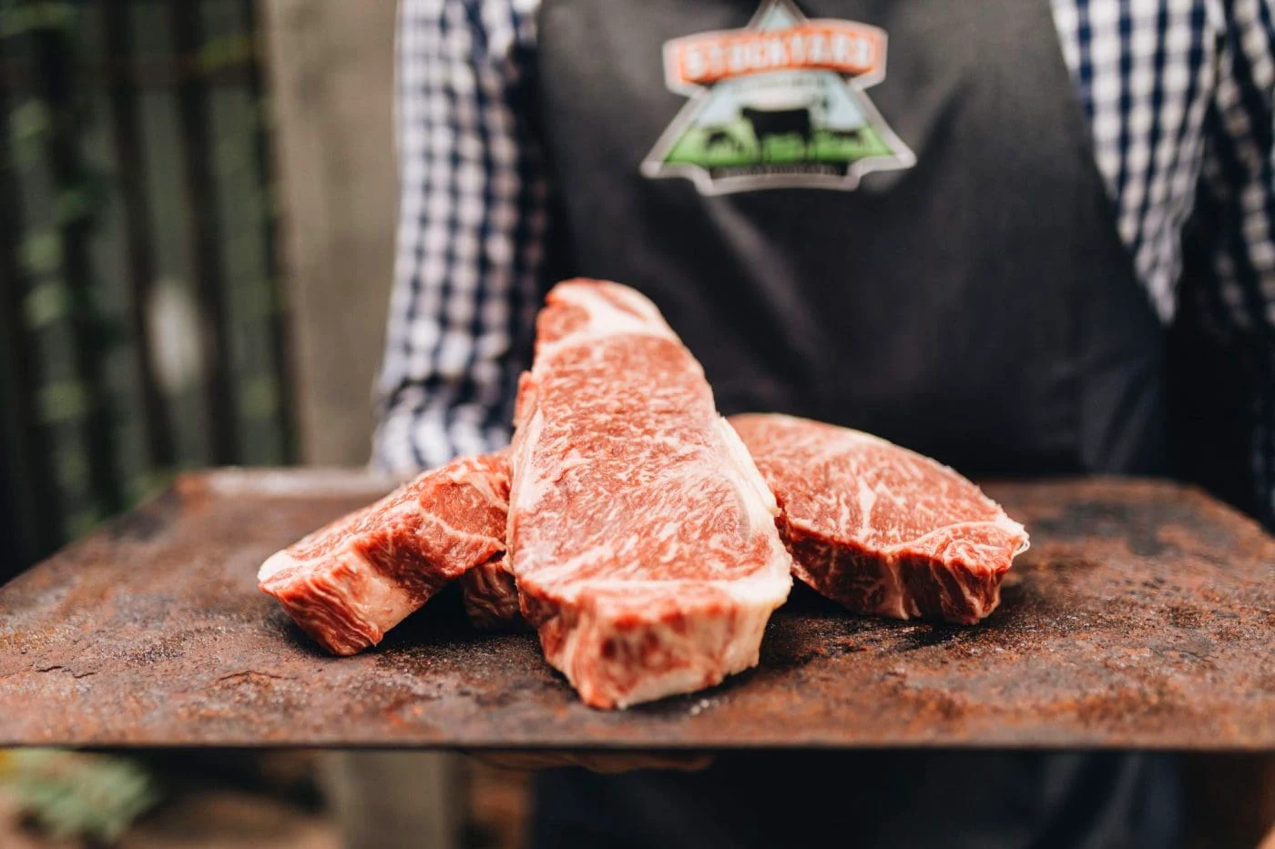 Stockyard butcher in a branded apron holding a wooden board with three premium grain-fed beef steaks
